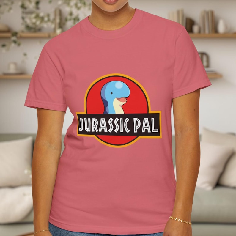 May include: A dusty rose short-sleeved t-shirt featuring a graphic of a blue dinosaur head inside a red circle with a yellow border. Below the circle, a black rectangular banner contains the white text JURASSIC PAL in a bold, sans-serif font.