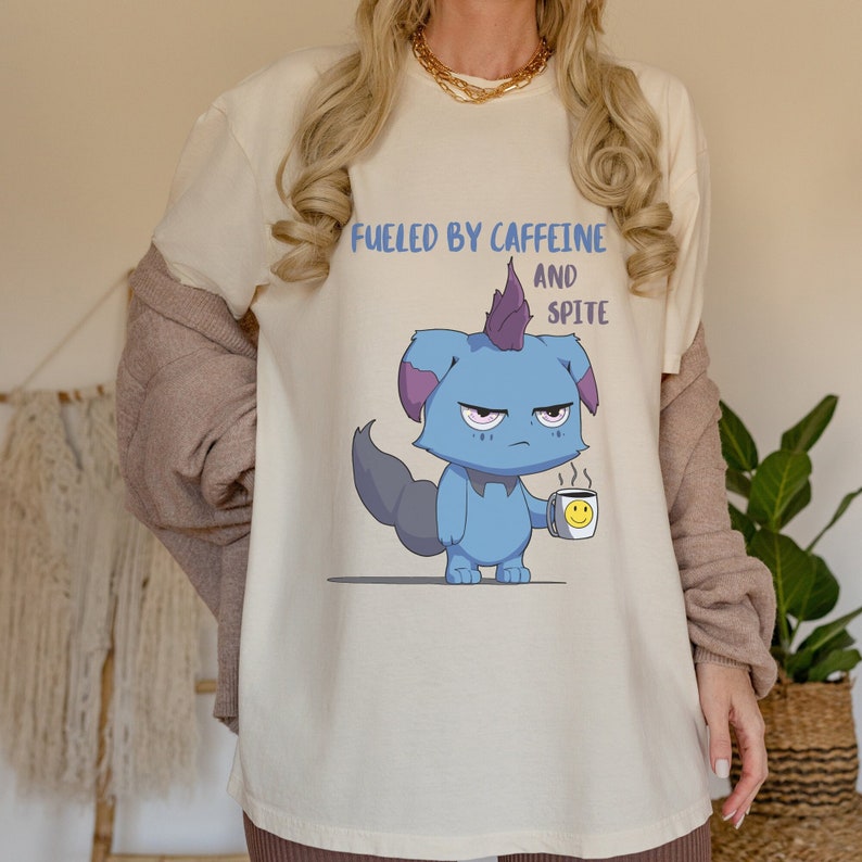 May include: Cream-colored t-shirt with the text "Fueled by Caffeine and Spite" above a cartoon blue creature holding a coffee mug with a smiley face. The creature has a purple mohawk and a grumpy expression.