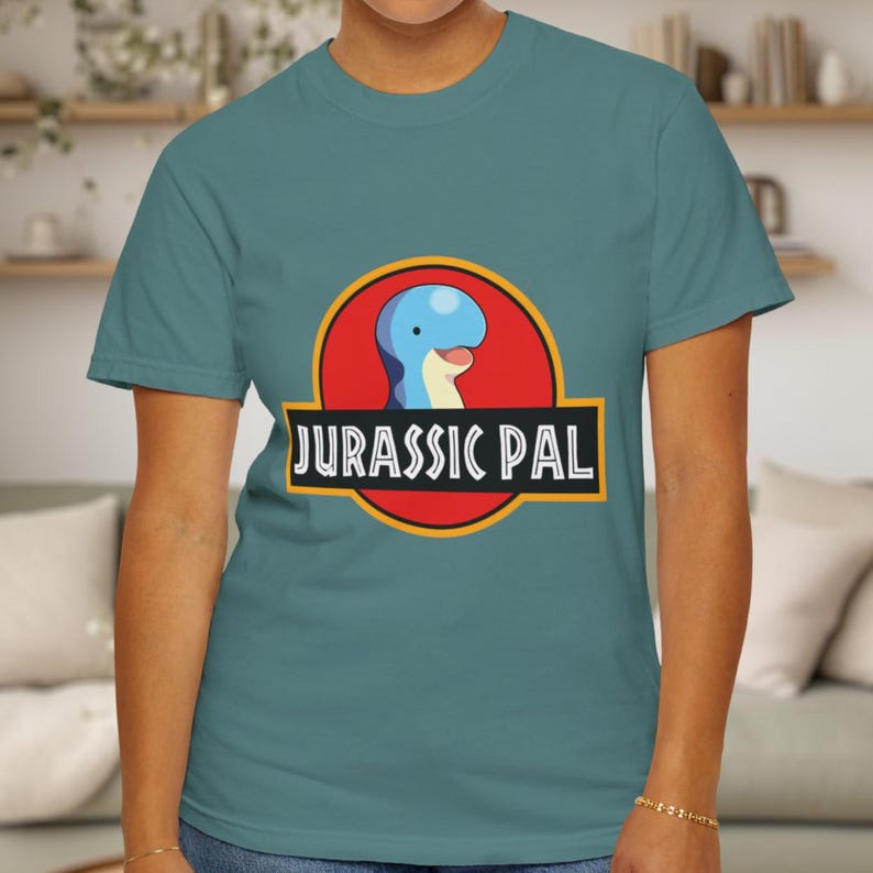 May include: A teal short-sleeve t-shirt featuring a graphic of a blue dinosaur head inside a red circle with a yellow border. Below the circle, a black rectangular banner contains the white text JURASSIC PAL in a bold, sans-serif font.