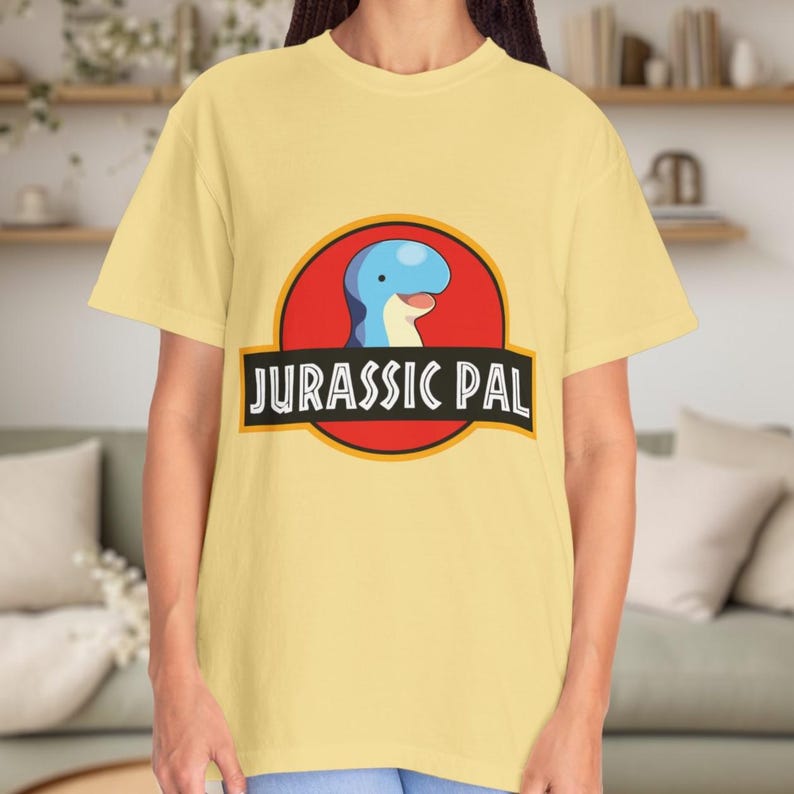 May include: A pale yellow short-sleeve t-shirt featuring a graphic of a blue dinosaur head inside a red circle with a yellow border. Below the circle, a black horizontal banner contains the white text JURASSIC PAL in a bold, sans-serif font.