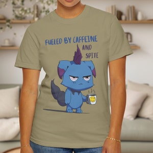 May include: A light olive green t-shirt featuring the phrase "FUELED BY CAFFEINE AND SPITE" above a cartoon creature. The blue creature holds a coffee mug with a smiley face.