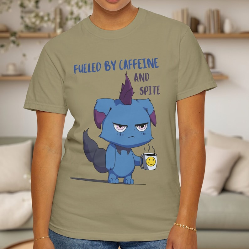May include: A light olive green t-shirt featuring the phrase "FUELED BY CAFFEINE AND SPITE" above a cartoon creature. The blue creature holds a coffee mug with a smiley face.