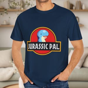 May include: A navy blue short-sleeved t-shirt featuring a circular graphic with a red background and a yellow border. The graphic displays a cartoon blue dinosaur head and the white text JURASSIC PAL centered within a black rectangular banner.