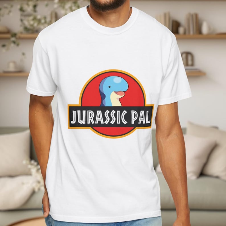 May include: A white short-sleeved t-shirt featuring a graphic of a blue cartoon dinosaur head inside a red circle with a yellow border. Below the circle is a black rectangular banner with the white text JURASSIC PAL printed in a bold font.