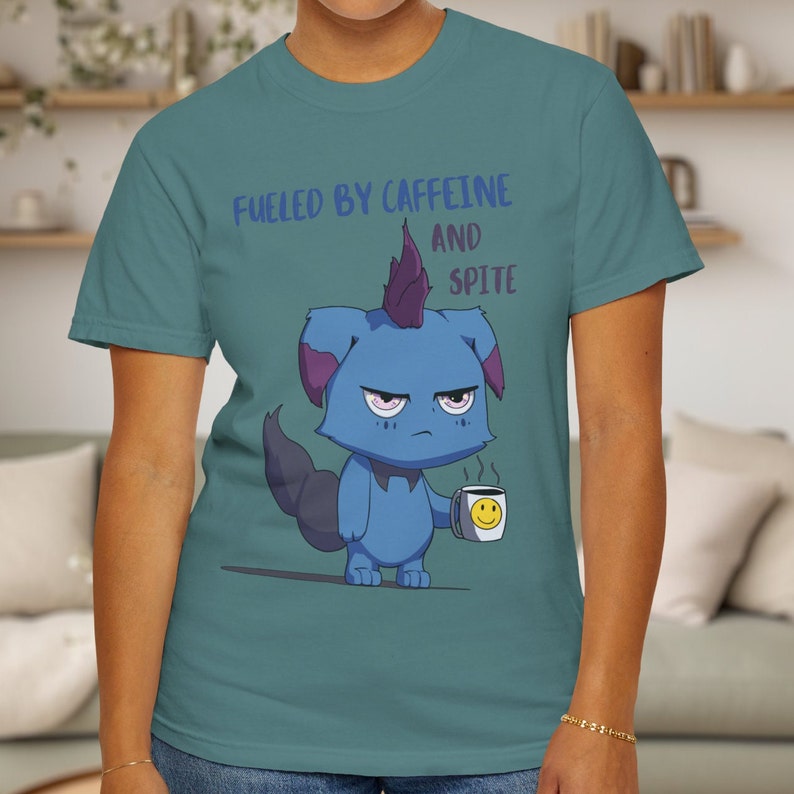 May include: Teal t-shirt with the text "Fueled by Caffeine and Spite." The shirt features a cartoon blue creature with a grumpy expression, holding a coffee mug with a smiley face.