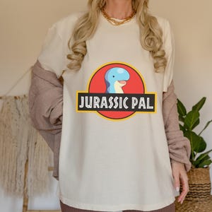 May include: A cream-colored short-sleeve t-shirt featuring a graphic of a blue cartoon dinosaur head inside a red circle with a yellow border. Below the circle, a black rectangular banner contains the white text JURASSIC PAL in a bold font.