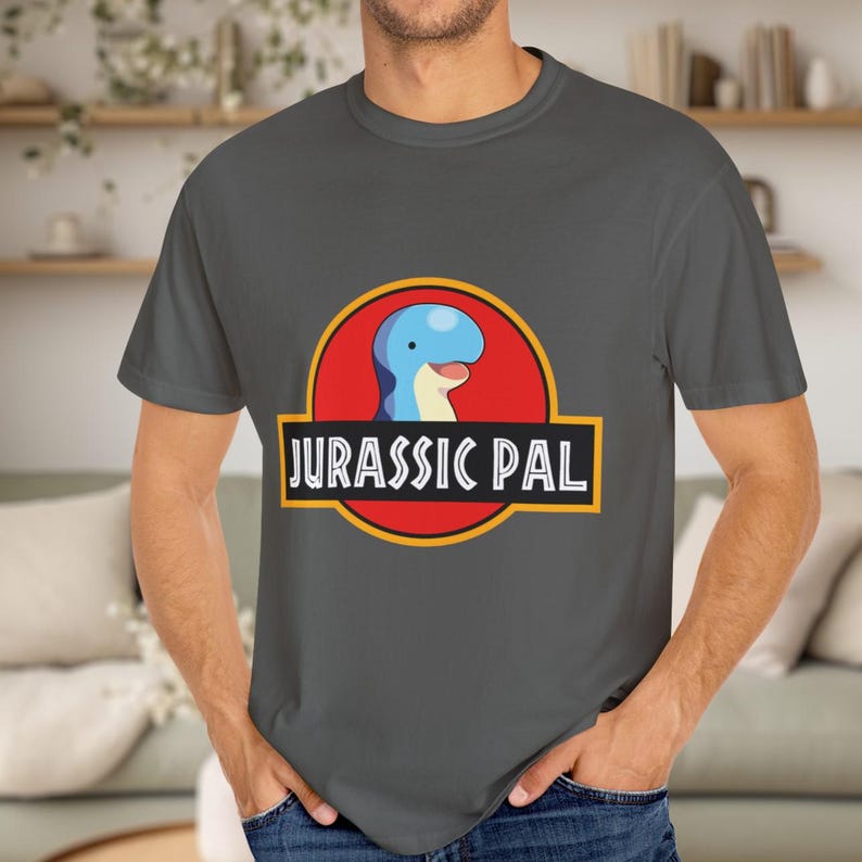May include: A dark gray short-sleeved t-shirt featuring a graphic of a blue and cream-colored cartoon dinosaur head inside a red circle with a yellow border. Below the circle is a black rectangular banner with the white text JURASSIC PAL.