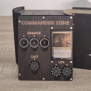 May include: A dark brown wooden Commander Zone game accessory with dials for damage, cast, and life. The text "COMMANDER ZONE" is at the top. A card is displayed on the right side.