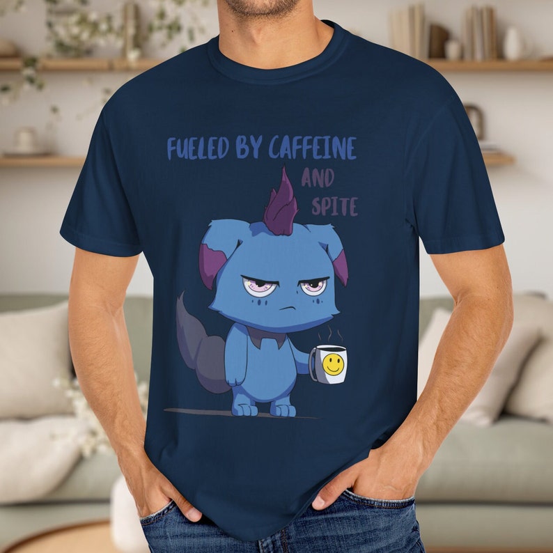 May include: Navy blue t-shirt featuring a grumpy blue cartoon creature with a purple mohawk holding a coffee mug with a smiley face. The text "FUELED BY CAFFEINE AND SPITE" is printed above the creature.
