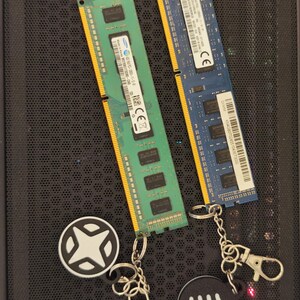 May include: Two RAM memory modules, one green and one blue, with gold connectors. Two keychains with round black and white charms are also present. The charms have a starburst design and a Wi-Fi symbol.