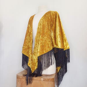 May include: A mustard yellow velvet shawl with black fringe trim. The shawl is open in the front and has a V-neckline.