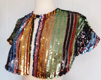 May include: A short-sleeved, rainbow sequined bolero with a keyhole neckline. The bolero is made of a sheer fabric with sequins in shades of blue, green, orange, red, and pink.