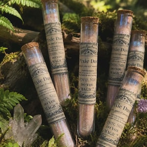 May include: Several clear glass test tubes filled with shimmering pink powder, each sealed with a cork stopper. A paper label on each tube reads Pixie Dust, includes instructions to Open, Stir, and asks the question DO YOU MAGIC? with Moon Apothecary branding.