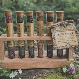 May include: A wooden test tube rack holding six glass tubes filled with various dried herbs and spices. Each tube has a cork stopper and a label with text. A tag with the words "moonflower apothecary herbal tea set" hangs from the rack.