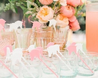 May include: A table setting with pink lemonade in a glass pitcher and clear glasses with pink and white unicorn stirrers. The table is decorated with a bouquet of pink and peach flowers.