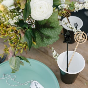 May include: A teal green plate with a silver chain and a white tag that says "AUGUST". The plate is on a wooden table with a white and black cup and a wooden stick with a black silhouette of a person holding a gun.