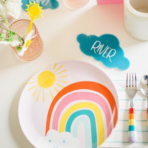 May include: A pink plate with a rainbow, sun, and cloud design. The plate is set on a white table with a blue cloud place card that says "River".