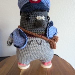 May include: A handmade crochet figure of a postal worker. The figure is gray with a blue hat and jacket, a brown satchel, and pink-soled shoes. The hat has a red emblem. The figure is standing on a wooden surface.