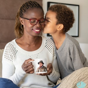 Peut inclure: Une tasse en c&eacute;ramique blanche avec une photo d'une femme portant un chapeau et le mot "VERA" imprim&eacute; au bas. Une jeune personne embrasse la personne tenant la tasse sur la joue.