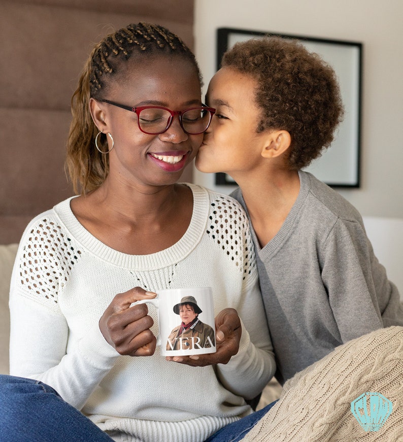 Peut inclure: Une tasse en c&eacute;ramique blanche avec une photo d'une femme portant un chapeau et le mot "VERA" imprim&eacute; au bas. Une jeune personne embrasse la personne tenant la tasse sur la joue.