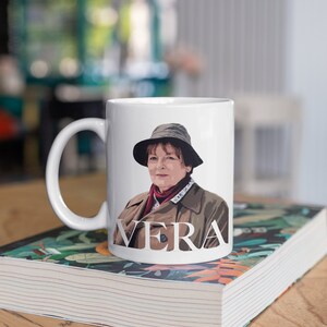 Peut inclure: Une tasse en c&eacute;ramique blanche avec une illustration en couleur d'une femme portant un chapeau et un manteau. La tasse porte l'inscription "VERA" sur le devant.