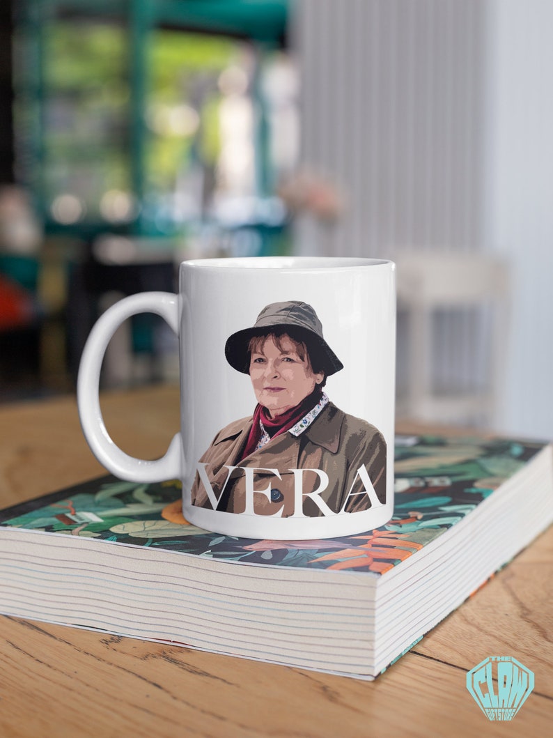 Peut inclure: Une tasse en c&eacute;ramique blanche avec une illustration en couleur d'une femme portant un chapeau et un manteau. La tasse porte l'inscription "VERA" sur le devant.