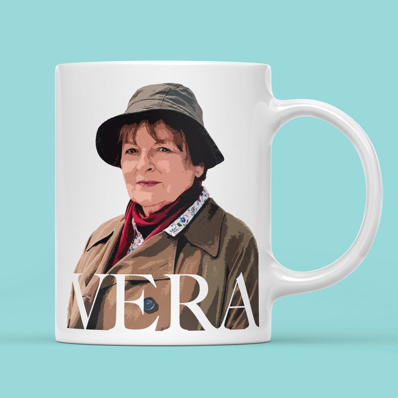 Peut inclure: Mug en c&eacute;ramique blanche avec une illustration en couleur d'une femme portant un chapeau et un manteau bruns. Le texte "VERA" est imprim&eacute; en blanc sur la tasse.