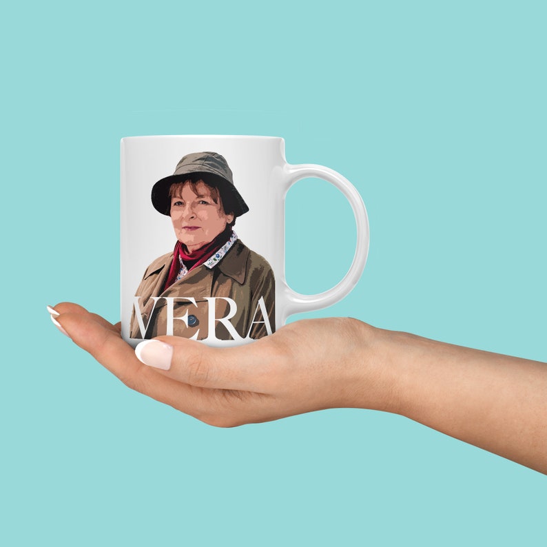 Peut inclure: Une tasse en c&eacute;ramique blanche avec une image couleur d'une femme portant un chapeau marron et une veste marron. Le texte "VERA" est imprim&eacute; en noir sur la tasse.