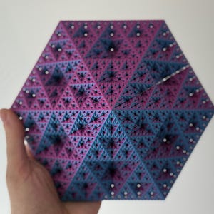 May include: A geometric, hexagonal art piece with a fractal design. The piece is held by a hand and features a complex pattern of triangles in shades of blue and pink, with small white dots.