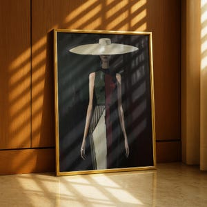 May include: A framed artwork featuring a fashion illustration of a person wearing a wide-brimmed hat and a color-blocked dress. The dress is divided into sections of green, red, and white stripes. The artwork is in a gold frame, leaning against a wooden wall.