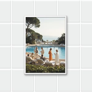 以下が含まれることがあります： フレームに入ったプリントは、プールサイドで交流する人々のヴィンテージシーンを描いています。この画像には、スタイリッシュな服装の人々、ヤシの木、海、澄み切った青い空を背景にした風景が写っています。全体的な美しさは、レジャーと優雅さを連想させます。