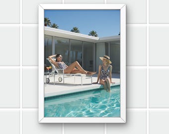 May include: A framed photograph shows two people in swimwear by a swimming pool. One is reclining on a sun lounger, the other is sitting on the pool's edge. The scene is set against a modern house and palm trees under a clear blue sky.