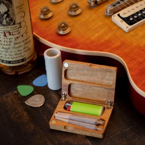 May include: A wooden box containing a lighter and two rolled cigarettes, a white ceramic tube, guitar plectrums, and a bottle of bourbon on a dark wooden surface. A sunburst electric guitar is partially visible in the background.