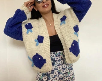 May include: A cream-colored, open-front cardigan with blue sleeves and blue floral appliqués. The cardigan is paired with a floral print skirt. The sleeves are a solid blue color, and the floral appliqués are a darker blue with light blue accents.
