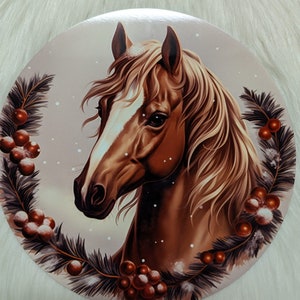May include: A circular decorative item with a detailed illustration of a brown horse head with a white blaze. The horse is framed by a wreath of pine needles and red berries, set against a soft, snowy background.