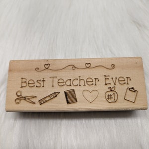 May include: Wooden stamp with the text "Best Teacher Ever" and five icons: scissors, crayon, notebook, heart, and an apple with the number one on it.