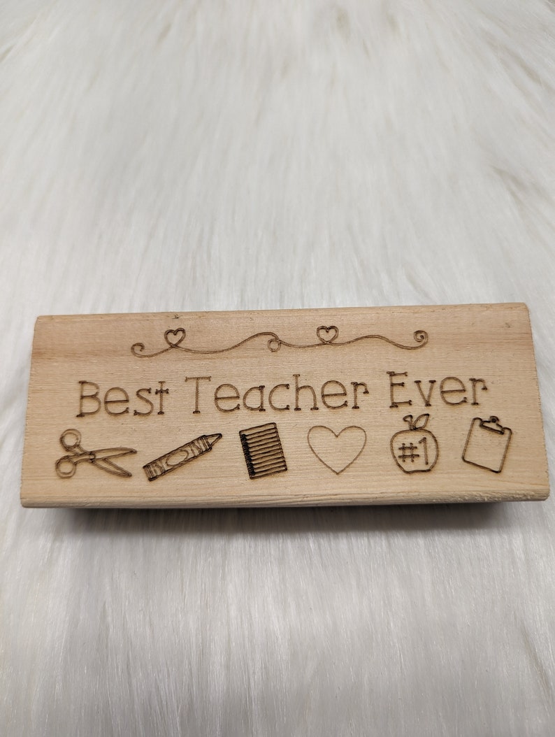 May include: Wooden stamp with the text "Best Teacher Ever" and five icons: scissors, crayon, notebook, heart, and an apple with the number one on it.