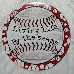 May include: A white and red round sign with a baseball graphic and the text "Living life by the seams".