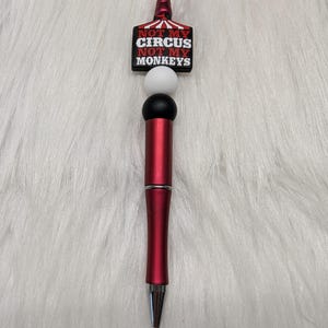 May include: A red ballpoint pen featuring a topper with a red and black circus tent design and the text NOT MY CIRCUS NOT MY MONKEYS in white lettering. The pen includes a white bead and a black bead positioned below the decorative topper.