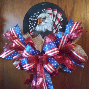 May include: A patriotic wreath with a red, white and blue bow and a round wooden sign featuring a painted bald eagle with American flags.