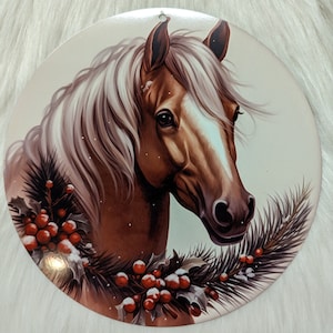 May include: A round Christmas bauble featuring a brown and white horse with a snowy evergreen branch and red berries.