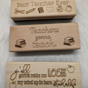 May include: Three wooden signs with engraved text and images. The top sign reads "Best Teacher Ever" with a pair of scissors, a crayon, a notebook, a heart, and a number one apple. The middle sign reads "Teachers gonna teach." with a stack of books. The bottom sign reads "Y'all gonna make me LOSE my mind up in here #teacherlife" with an apple.