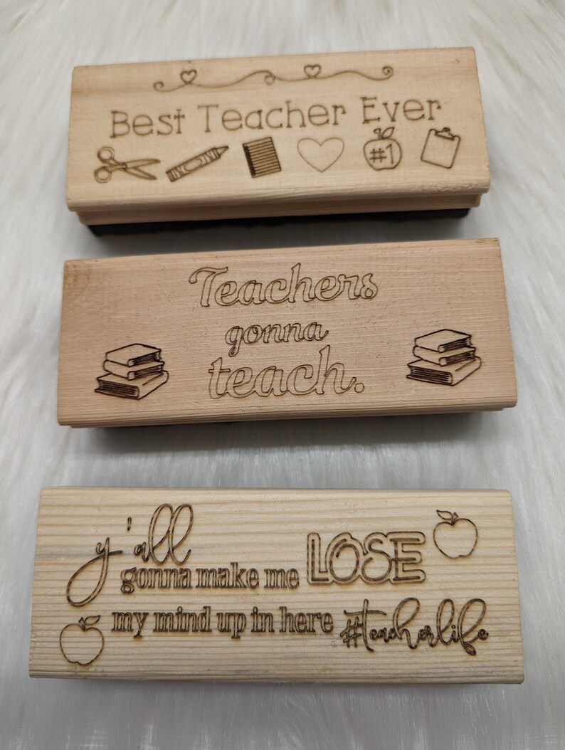 May include: Three wooden signs with engraved text and images. The top sign reads "Best Teacher Ever" with a pair of scissors, a crayon, a notebook, a heart, and a number one apple. The middle sign reads "Teachers gonna teach." with a stack of books. The bottom sign reads "Y'all gonna make me LOSE my mind up in here #teacherlife" with an apple.