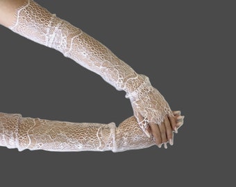 Könnte beinhalten: Ein Paar fingerlose Handschuhe aus weißer Spitze, die bis zum Oberarm reichen. Die Handschuhe weisen ein kompliziertes Blumenmuster mit gebogten Rändern oben und unten auf und bestehen aus einem zarten, halbtransparenten Stoff, der Unterarm und Hand bedeckt.