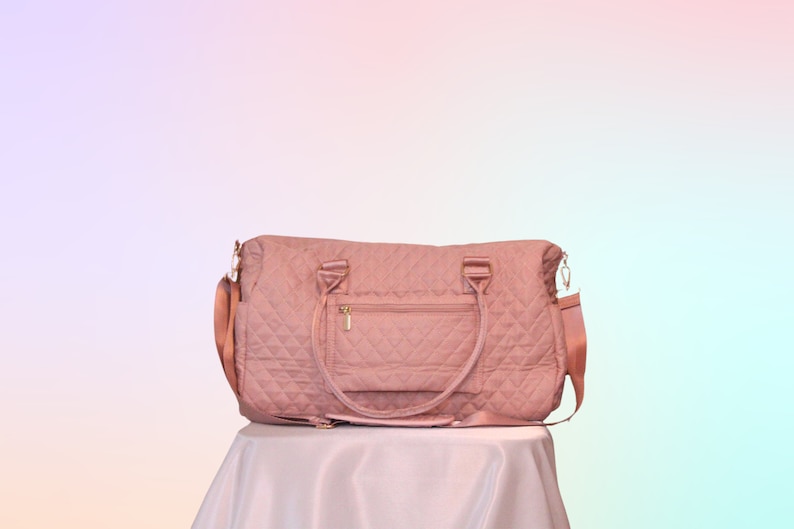 May include: A quilted pink travel bag with a gold zipper and a small pocket on the front. The bag has two top handles and a long adjustable strap.