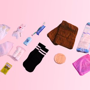 May include: A collection of baby essentials including diapers, wipes, a knitted bunny toy, socks, a brown towel, a pink bottle, a small yellow box, and a wooden tag with the text "It's a baby".