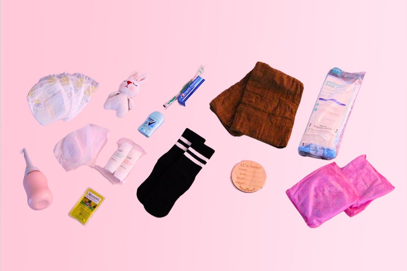 May include: A collection of baby essentials including diapers, wipes, a knitted bunny toy, socks, a brown towel, a pink bottle, a small yellow box, and a wooden tag with the text "It's a baby".