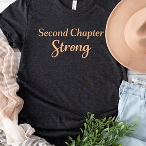 Può includere: T-shirt grigio scuro con la scritta "Second Chapter Strong" in corsivo arancione chiaro. Un cappello color cachi, jeans azzurri e un rametto di fogliame verde sono disposti intorno alla maglietta. È visibile anche una sciarpa a righe.