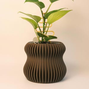 May include: A brown, ribbed plant pot with a green leafy plant and a yellow plant support. The pot has a unique, curved shape. The plant has large, heart-shaped leaves. The background is a solid, light beige colour.