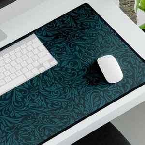 May include: A teal and black patterned desk mat with a white keyboard and a white mouse on top. The desk mat is on a white desk with a computer monitor in the background.